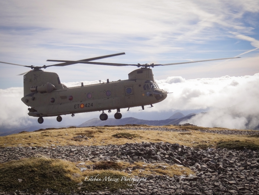 Un Chinook tomando en la cresta cresta de una montaña por encima del techo de nubes.